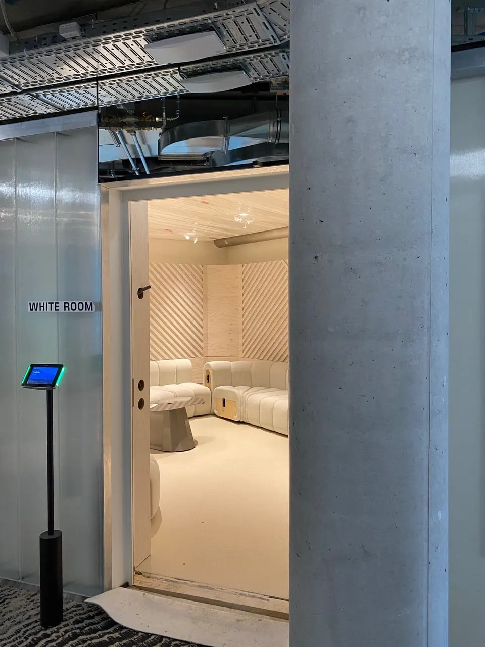 Eingang zum White Room neben einer Betonstütze, dahinter die helle Sitzlandschaft